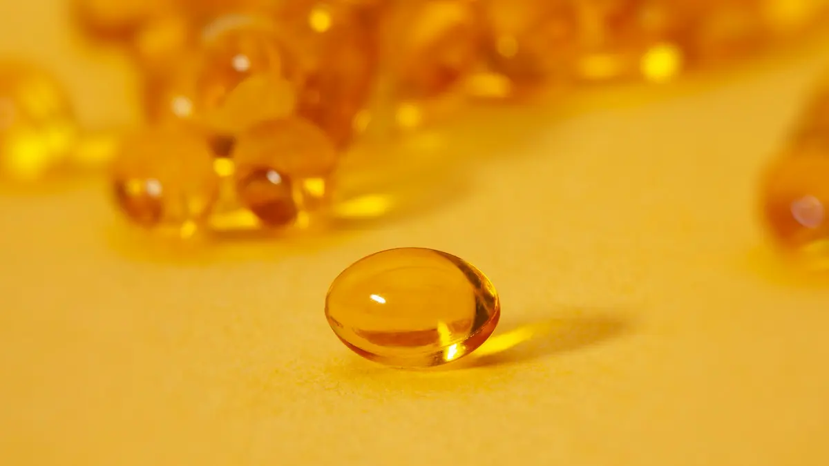 A single golden softgel capsule in front of a blurred pile of capsules on a yellow surface