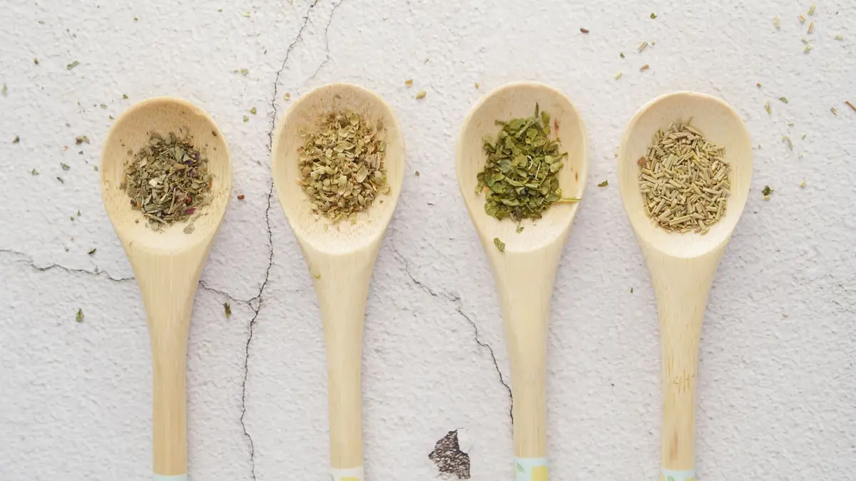 Four wooden spoons holding different dried herbs on a light background