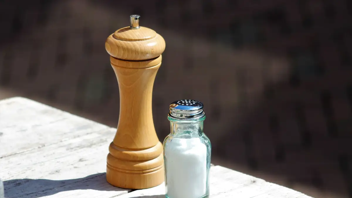 A glass salt shaker next to a wooden pepper mill on a sunlit wooden table