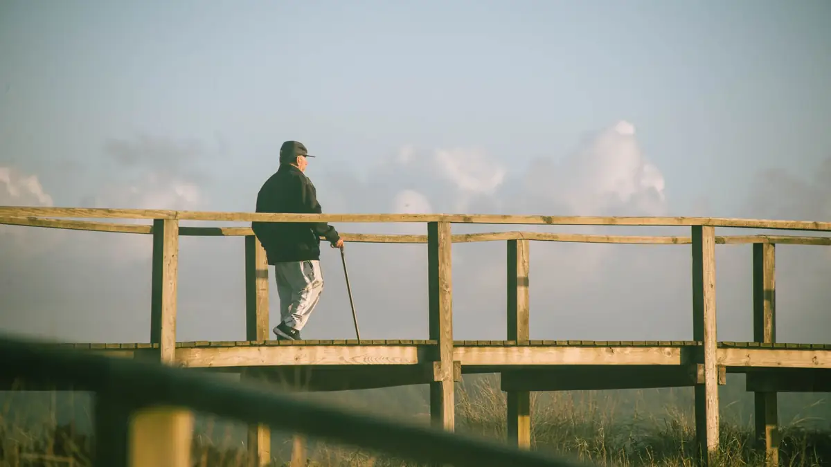 Older man with a walking stick strolling along a wooden boardwalk under a cloudy sky