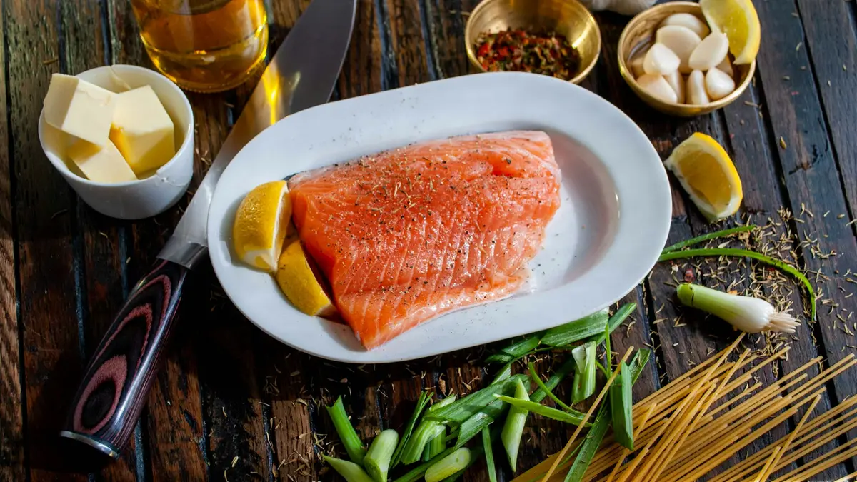 Raw salmon fillet on a white plate with lemon wedges, butter, garlic, and herbs on a wooden table