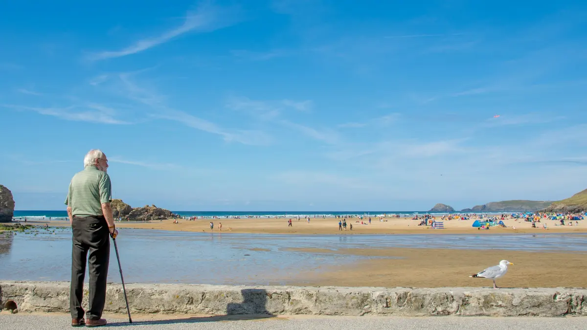 Older man with a cane standing in the sun, looking out over a beach under a blue sky